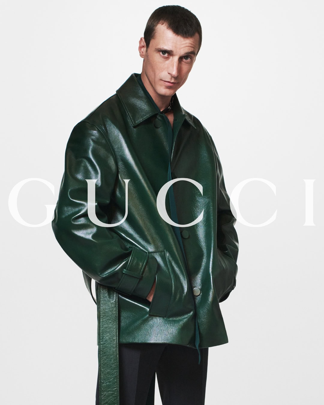 A fashionable adult male poses against a white backdrop wearing a green patent leather jacket and black pants. Brand logo visible.