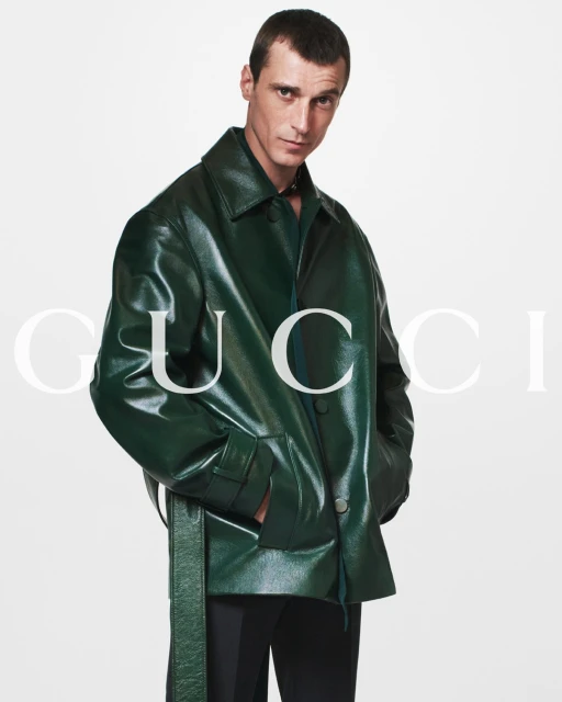 A fashionable adult male poses against a white backdrop wearing a green patent leather jacket and black pants. Brand logo visible.