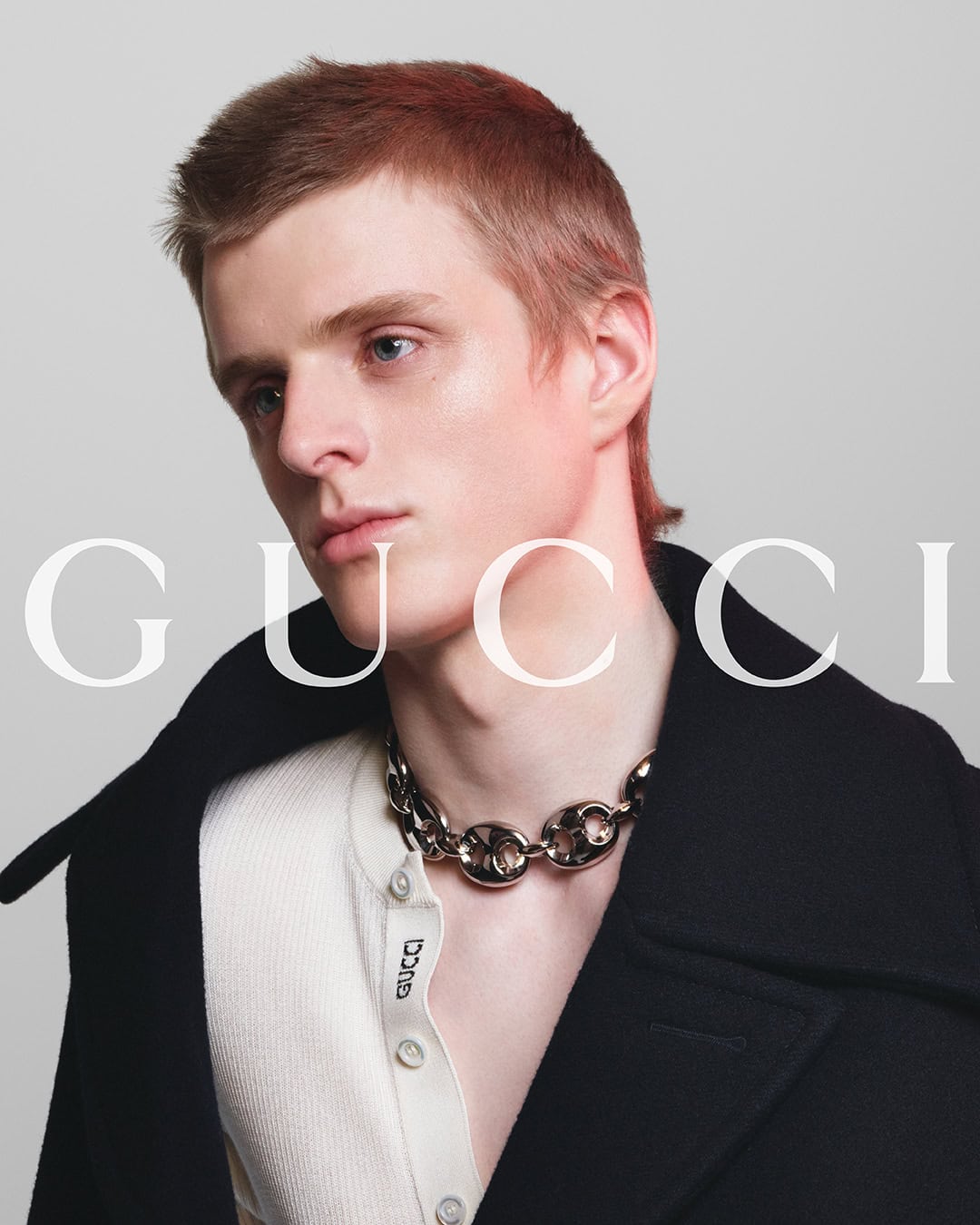 Close-up of a red-haired man in a black jacket and silver necklace with the word GUCCI in the foreground. Fashion advertisement.