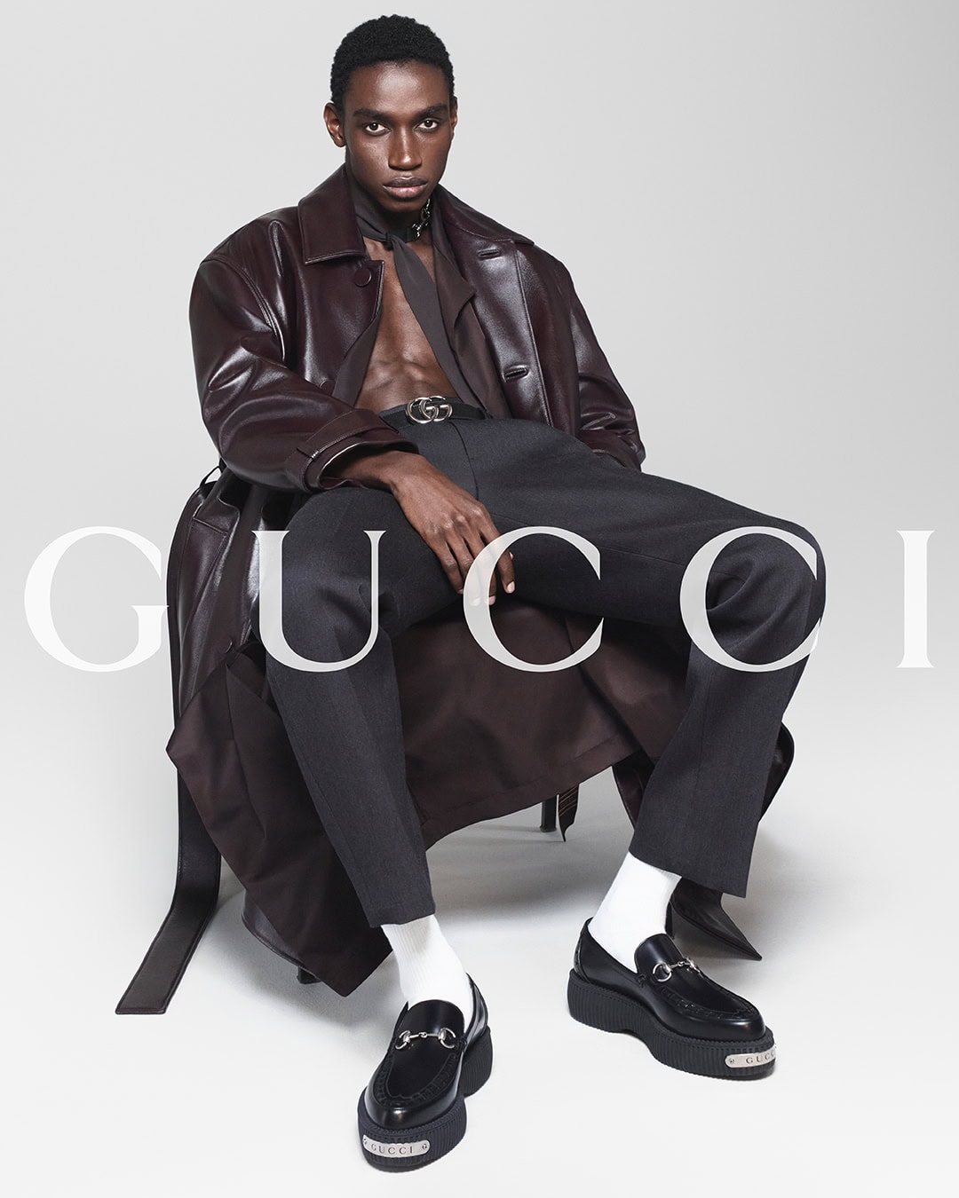 A dark-skinned male model sits in a studio wearing a brown leather trench coat, black pants, white socks, and black loafers, with Gucci text overlay.