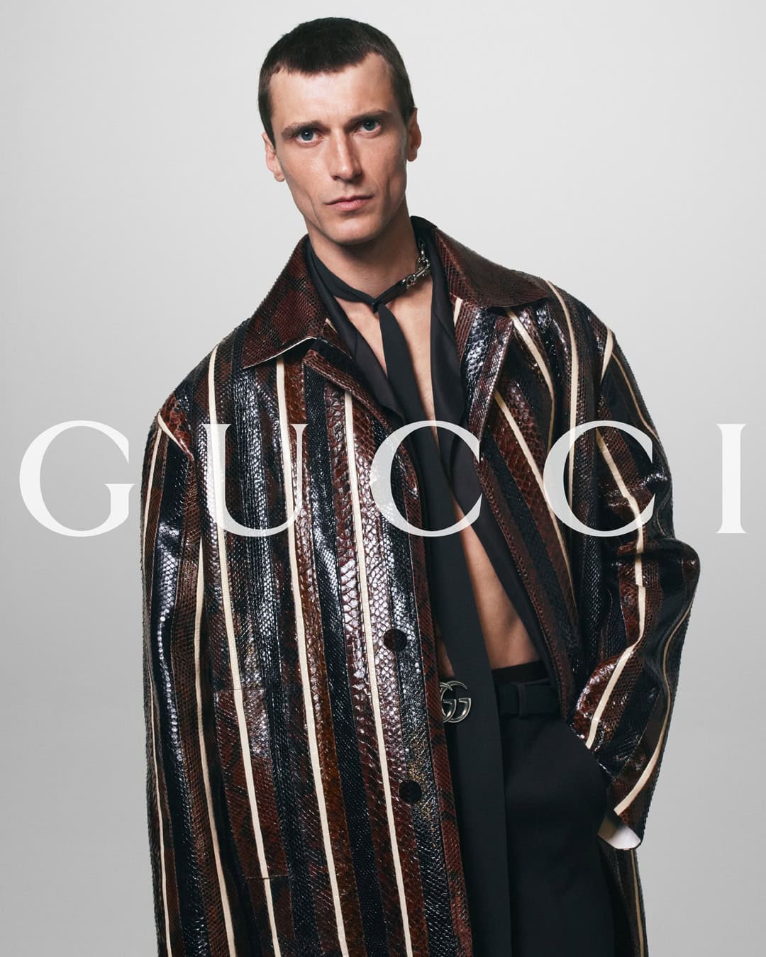 Man in Gucci striped coat, black shirt, and pants posing indoors. Fashion editorial shot on white background with brand logo.