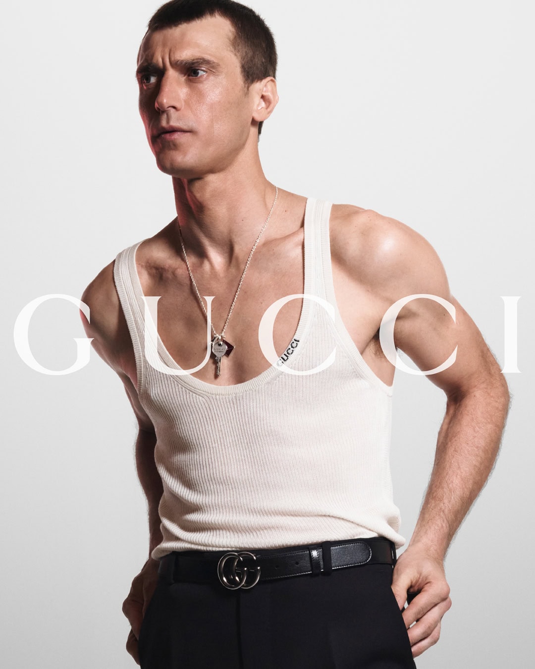 Portrait of a male fashion model wearing a white tank top and black pants with a Gucci belt against a white backdrop.