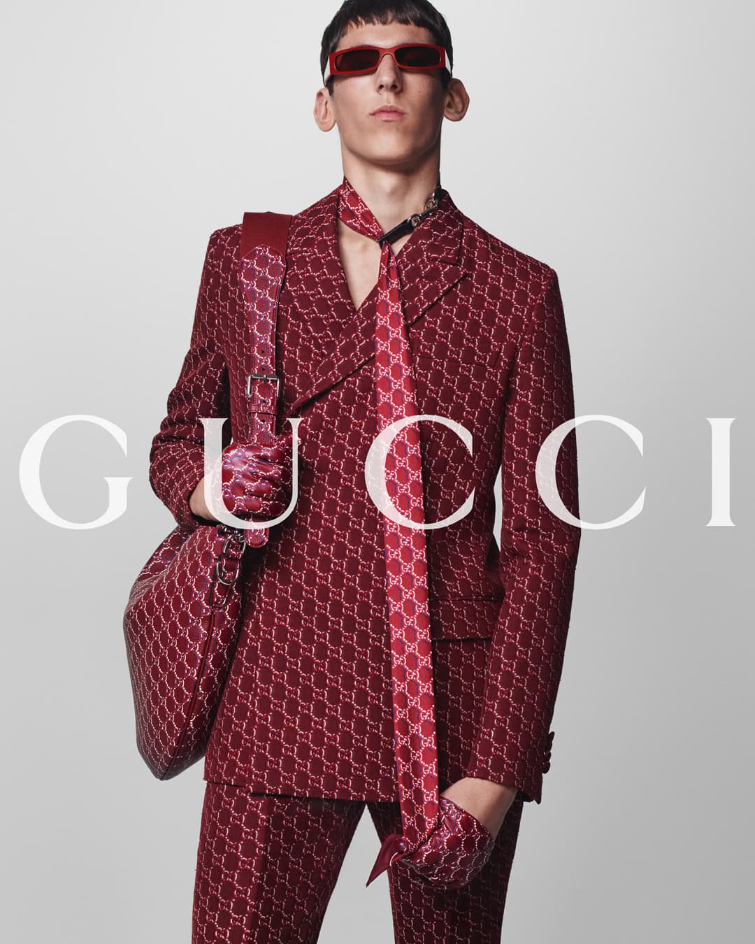 Man wearing red patterned suit and matching red sunglasses carrying a large shoulder bag against a white background
