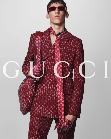Man wearing red patterned suit and matching red sunglasses carrying a large shoulder bag against a white background