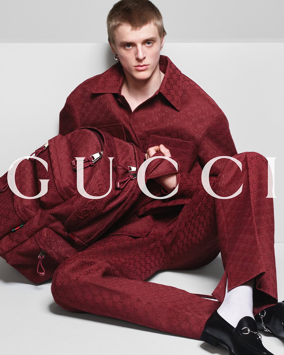A young adult male model poses with a designer backpack in a studio, wearing a patterned maroon suit, white socks, and black loafers.