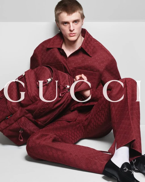 A young adult male model poses with a designer backpack in a studio, wearing a patterned maroon suit, white socks, and black loafers.