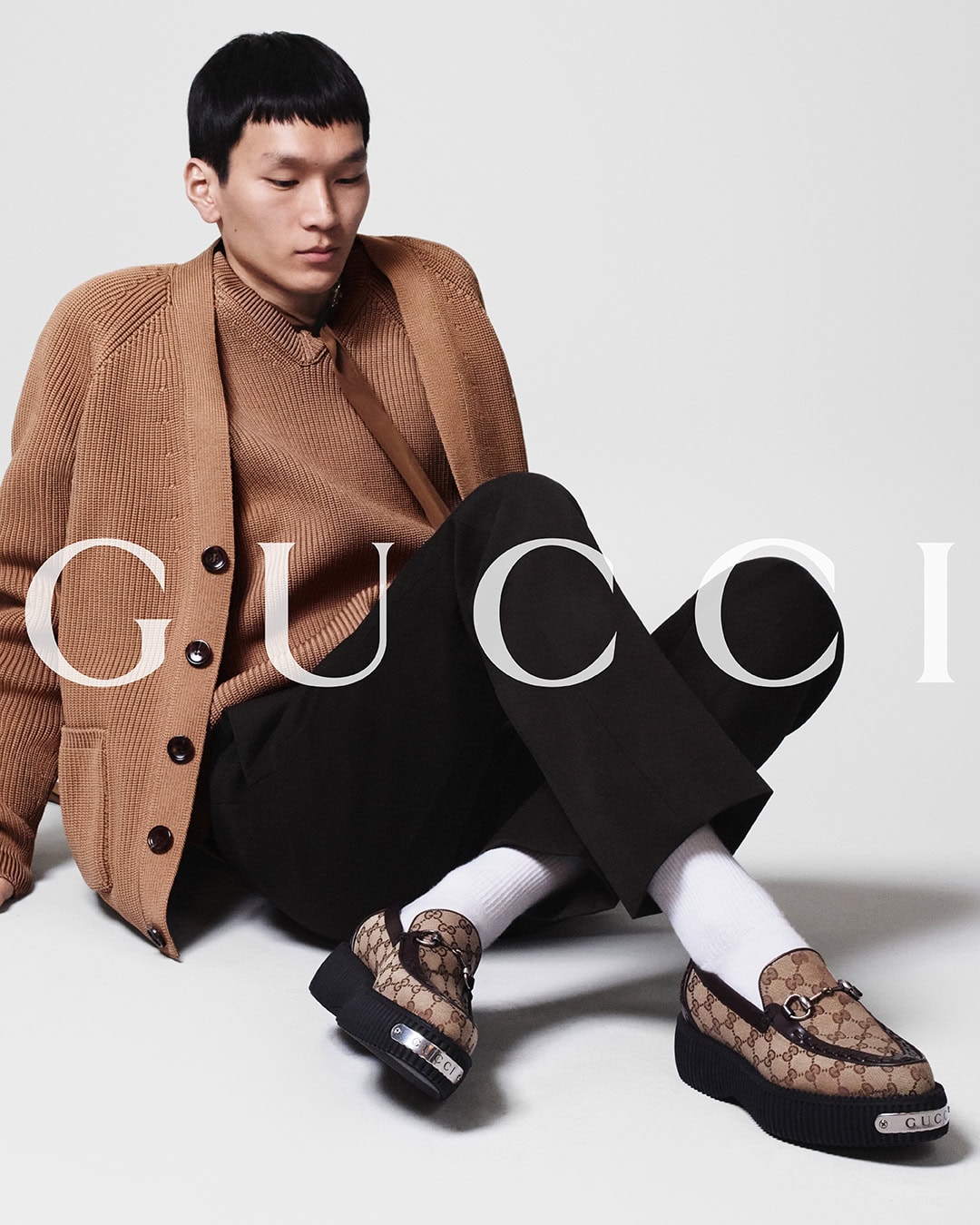 Asian man models Gucci loafers in a studio setting. He is wearing a tan cardigan, sweater, and black pants. Fashion and footwear.
