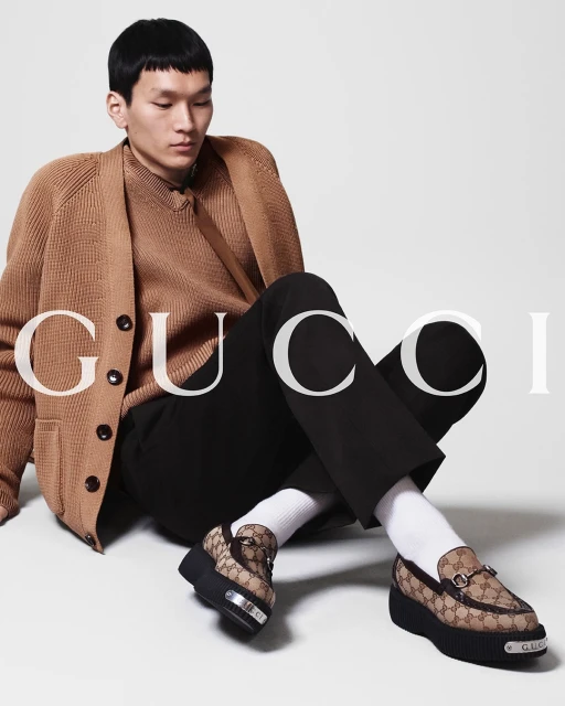 Asian man models Gucci loafers in a studio setting. He is wearing a tan cardigan, sweater, and black pants. Fashion and footwear.