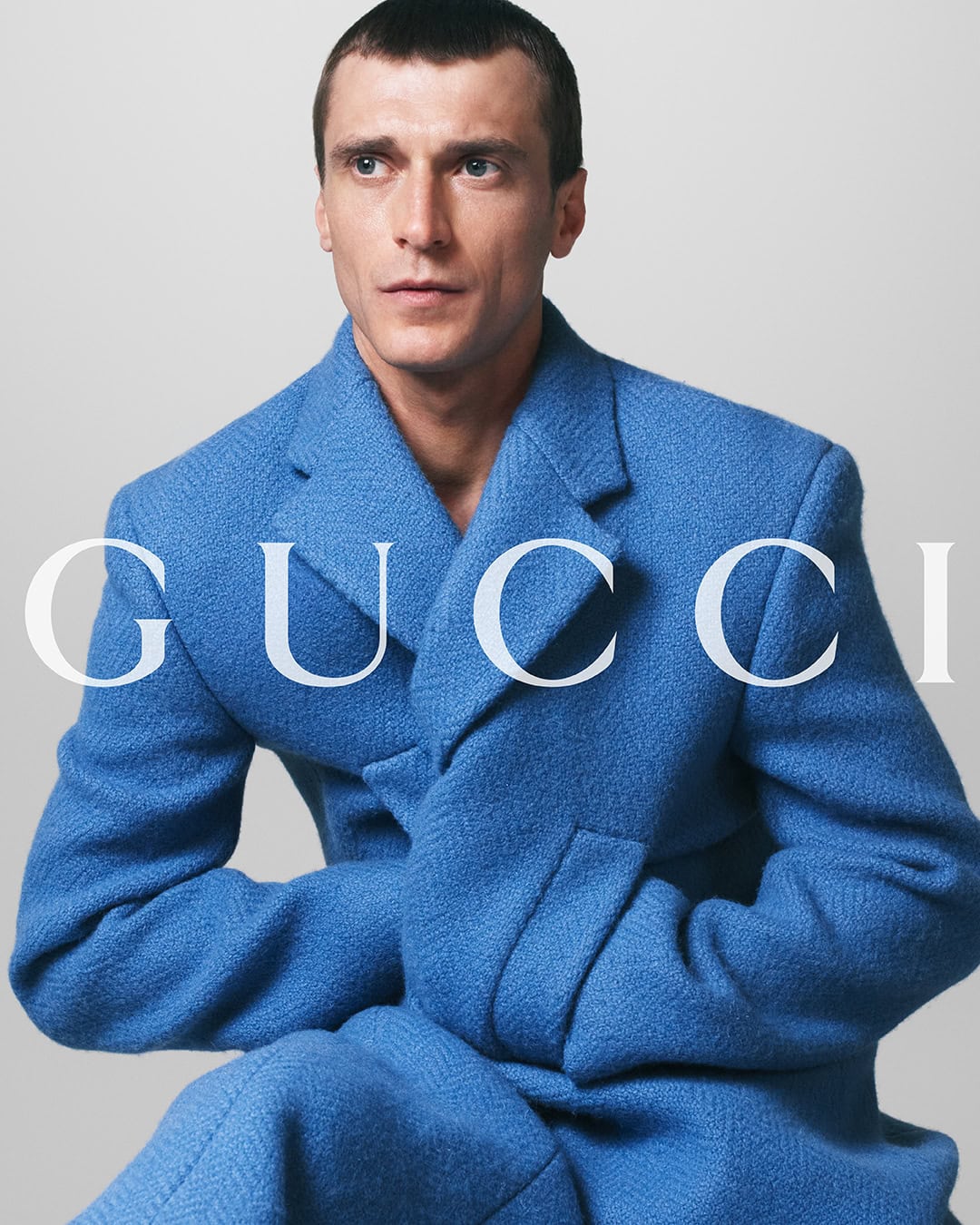 Portrait of a man in a blue textured double-breasted wool coat with the brand logo GUCCI superimposed on the front. Studio shot.