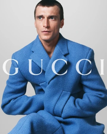 Portrait of a man in a blue textured double-breasted wool coat with the brand logo GUCCI superimposed on the front. Studio shot.