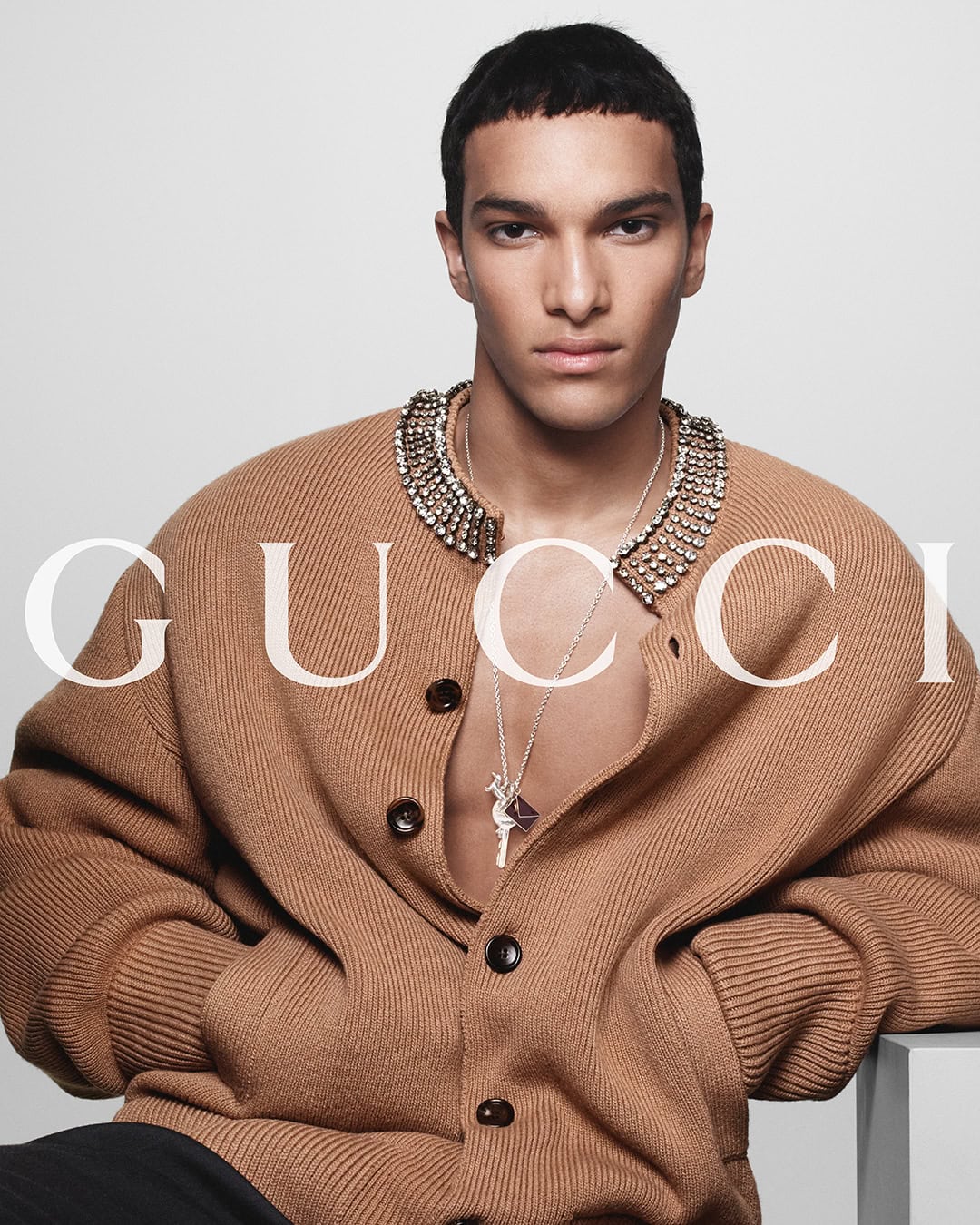 Fashion portrait of a young adult male in a tan cardigan with a jeweled collar and silver necklace, posing against a white backdrop.