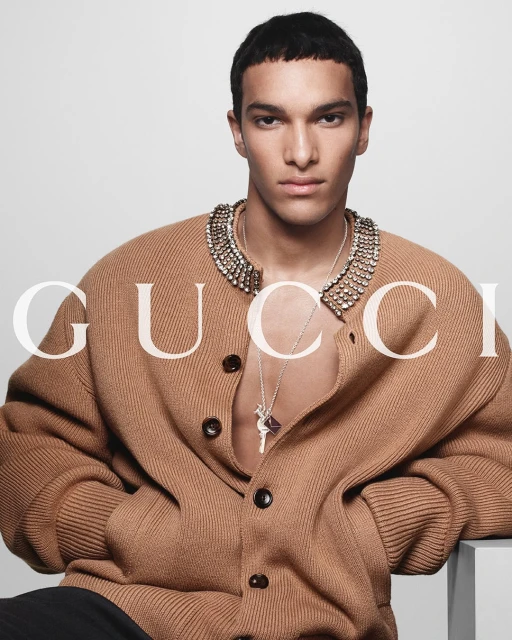 Fashion portrait of a young adult male in a tan cardigan with a jeweled collar and silver necklace, posing against a white backdrop.