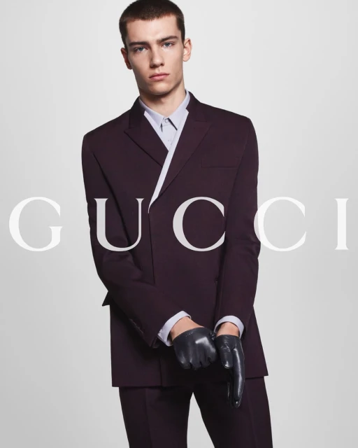 Young adult man in dark suit and gloves posing in a studio setting, featuring a stylish fashion portrait for an editorial or advertisement.