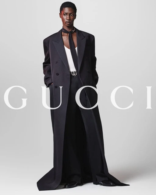 Fashion shot of an adult Black woman in a long black coat, white top, and black scarf, standing against a white background with brand text.