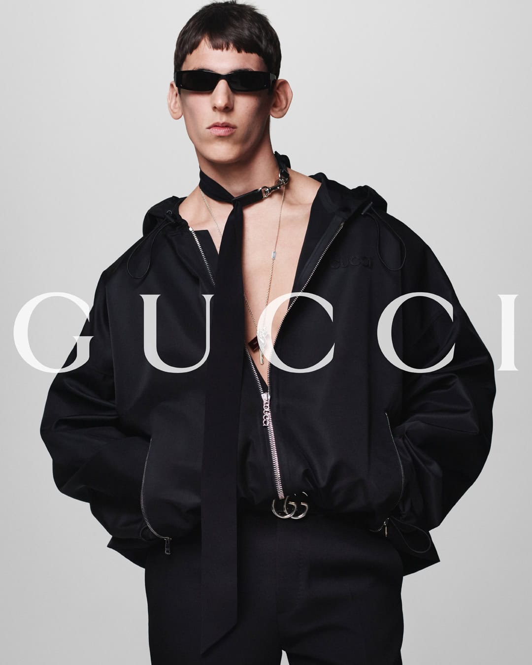 Fashion shot of a slim adult male wearing sunglasses and a black hooded jacket, posed against a light grey background.