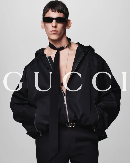 Fashion shot of a slim adult male wearing sunglasses and a black hooded jacket, posed against a light grey background.