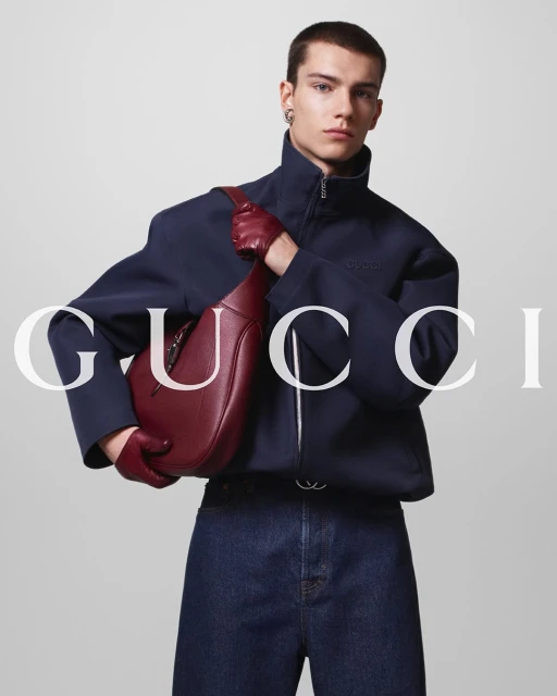 Male fashion model holds a burgundy leather Gucci bag, wearing a navy jacket, jeans, and gloves against a neutral backdrop.