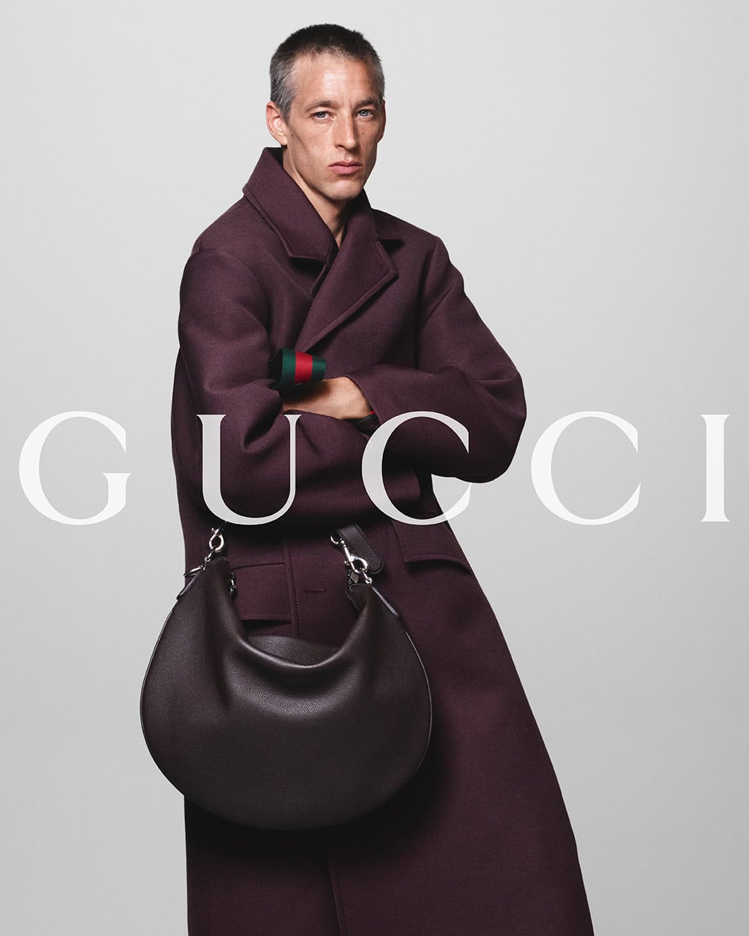 A man in a long, maroon coat holds a brown leather shoulder bag in front of the 'GUCCI' logo on a plain, light gray background.