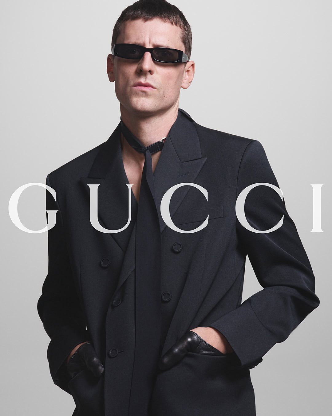 A man wearing a black Gucci suit, sunglasses, and gloves poses against a neutral gray studio backdrop in a fashion campaign.