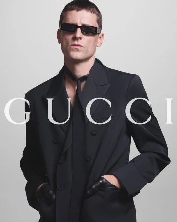 A man wearing a black Gucci suit, sunglasses, and gloves poses against a neutral gray studio backdrop in a fashion campaign.