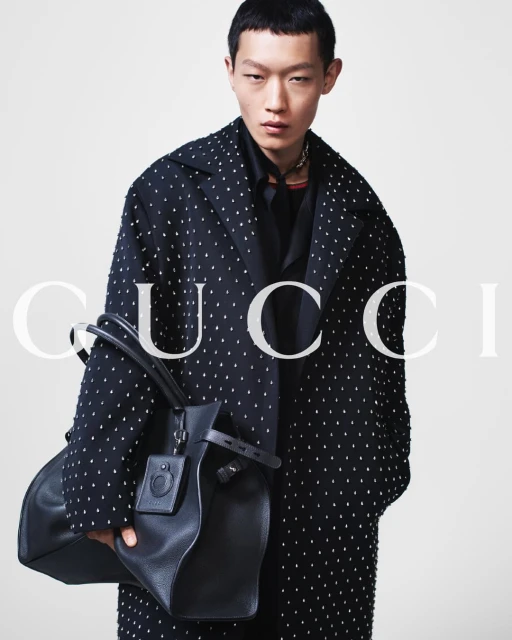 A fashionable Asian male model in a studded coat holds a black leather handbag in a studio setting with bright, soft lighting.