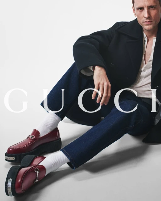 A man poses in dark denim jeans, a black peacoat, white socks, and burgundy leather loafers by Gucci on a white background.
