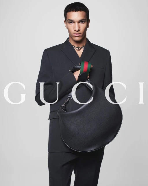 A man in a dark suit poses indoors holding a black leather bag with the Gucci logo visible in the background, fashion advertisement.