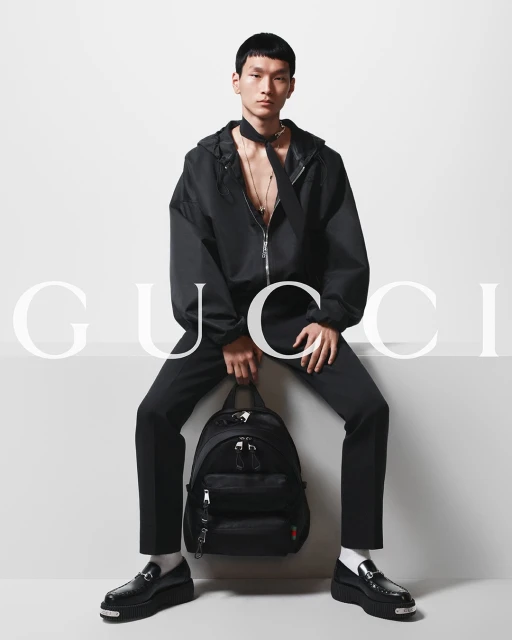 Asian man poses in studio wearing black jacket, pants and platform loafers, holding a Gucci backpack, with logo in background.