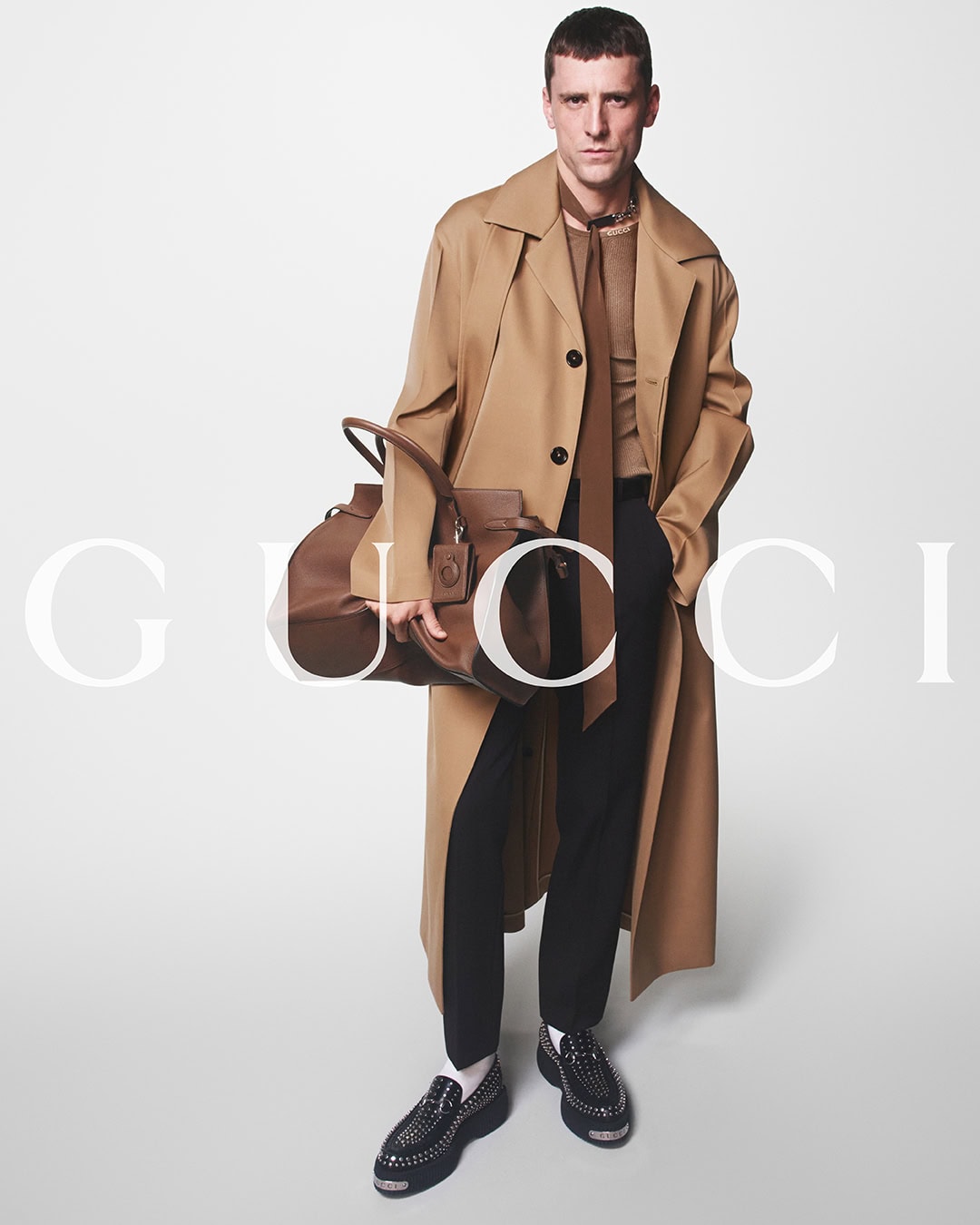 A stylish adult male model poses in a tan trench coat, brown shirt and tie, and black pants holding a leather duffel bag in a studio.