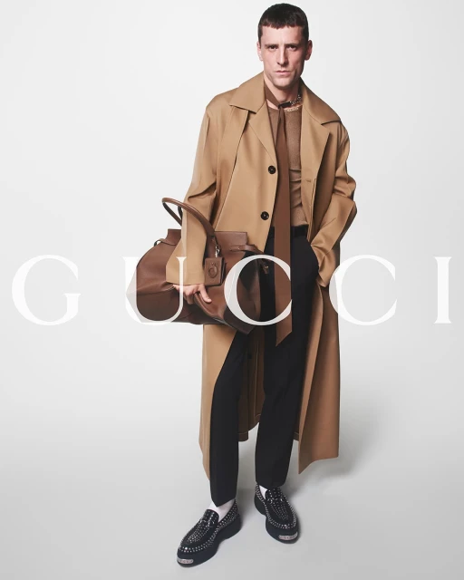 A stylish adult male model poses in a tan trench coat, brown shirt and tie, and black pants holding a leather duffel bag in a studio.