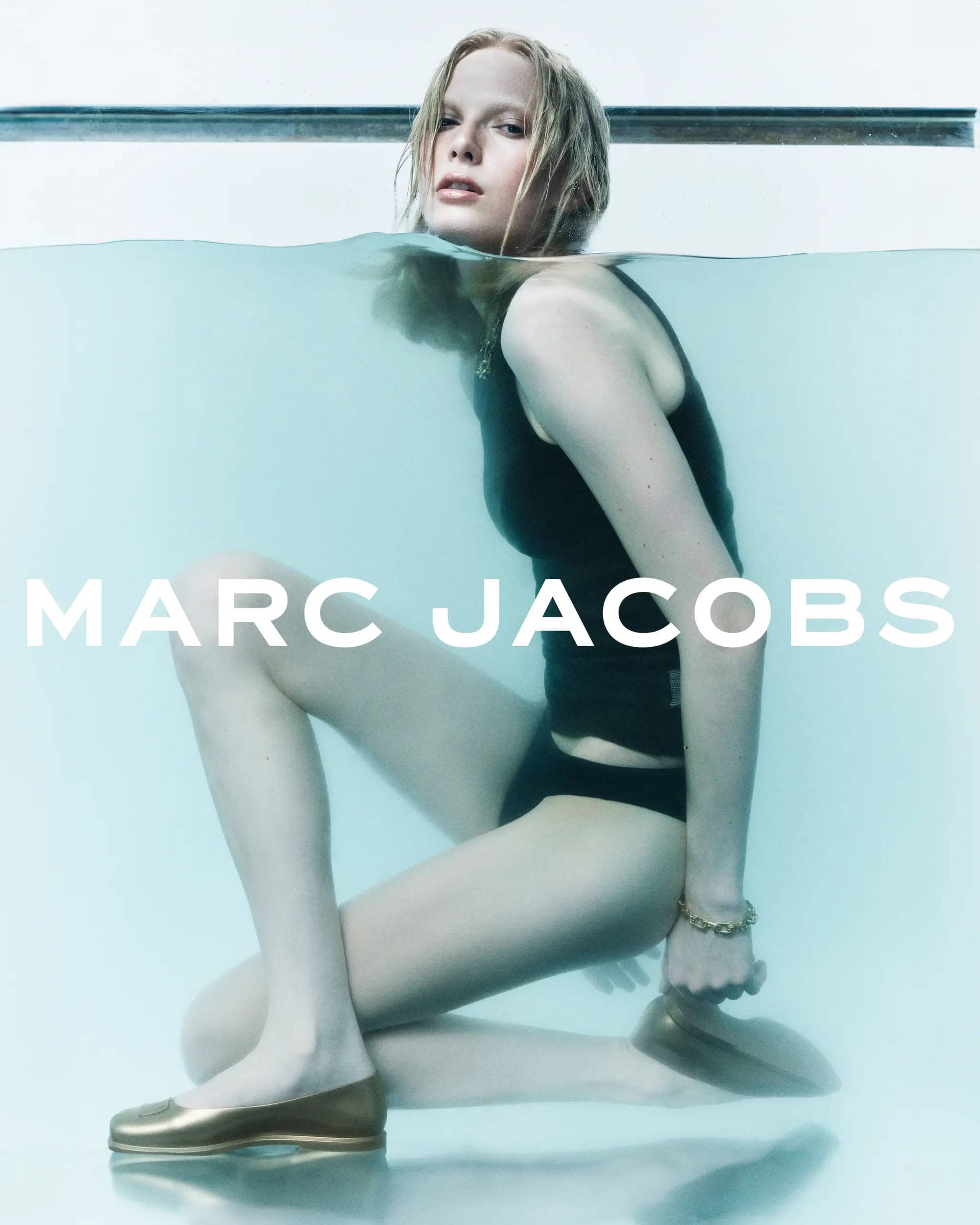 A fair-skinned woman in a black swimsuit and gold flats sits underwater in a bright pool wearing Marc Jacobs jewelry, a fashion campaign.