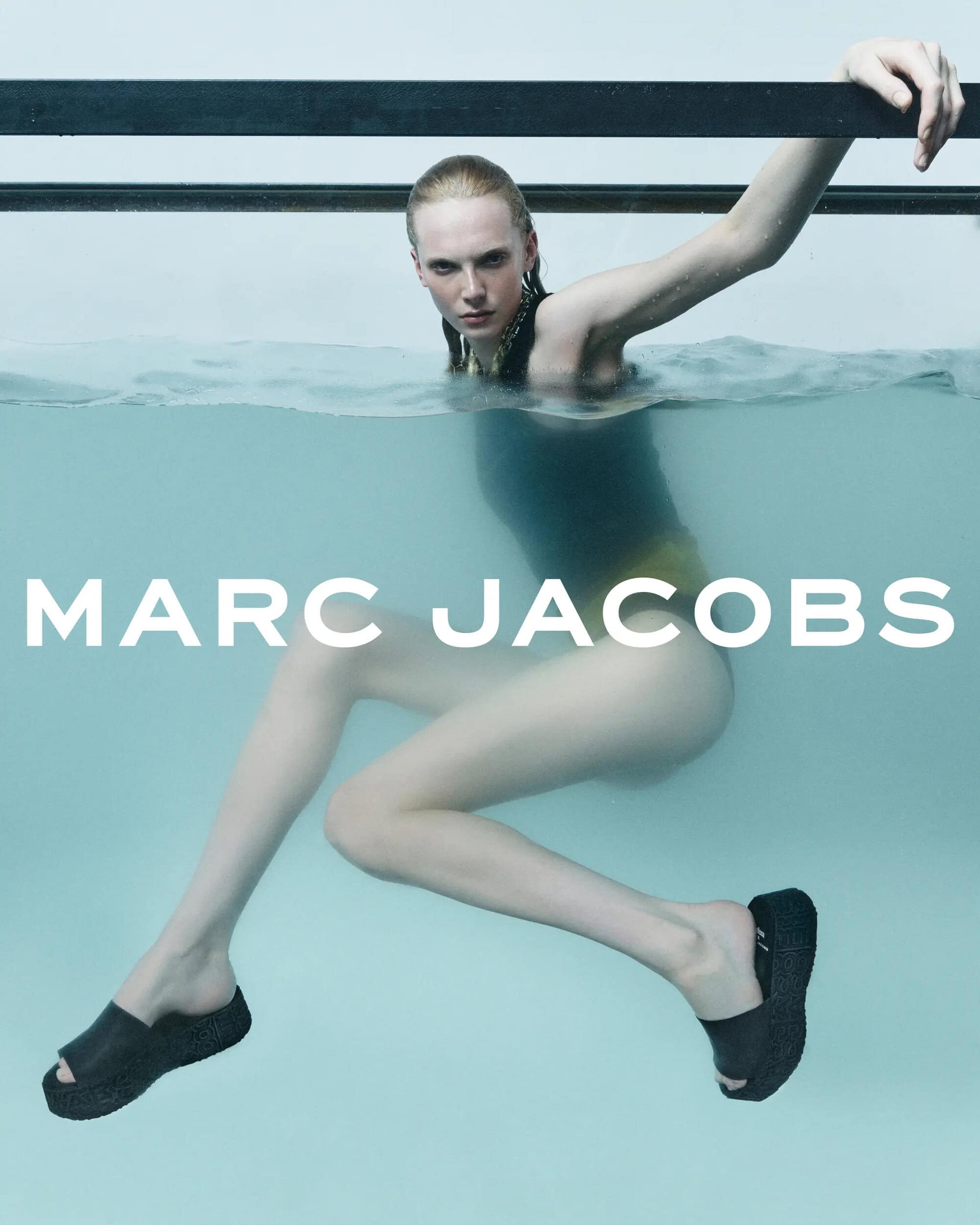 Fashion shot of a woman in the water wearing a swimsuit and platform sandals, leaning on a metal rail. Marc Jacobs logo is visible.