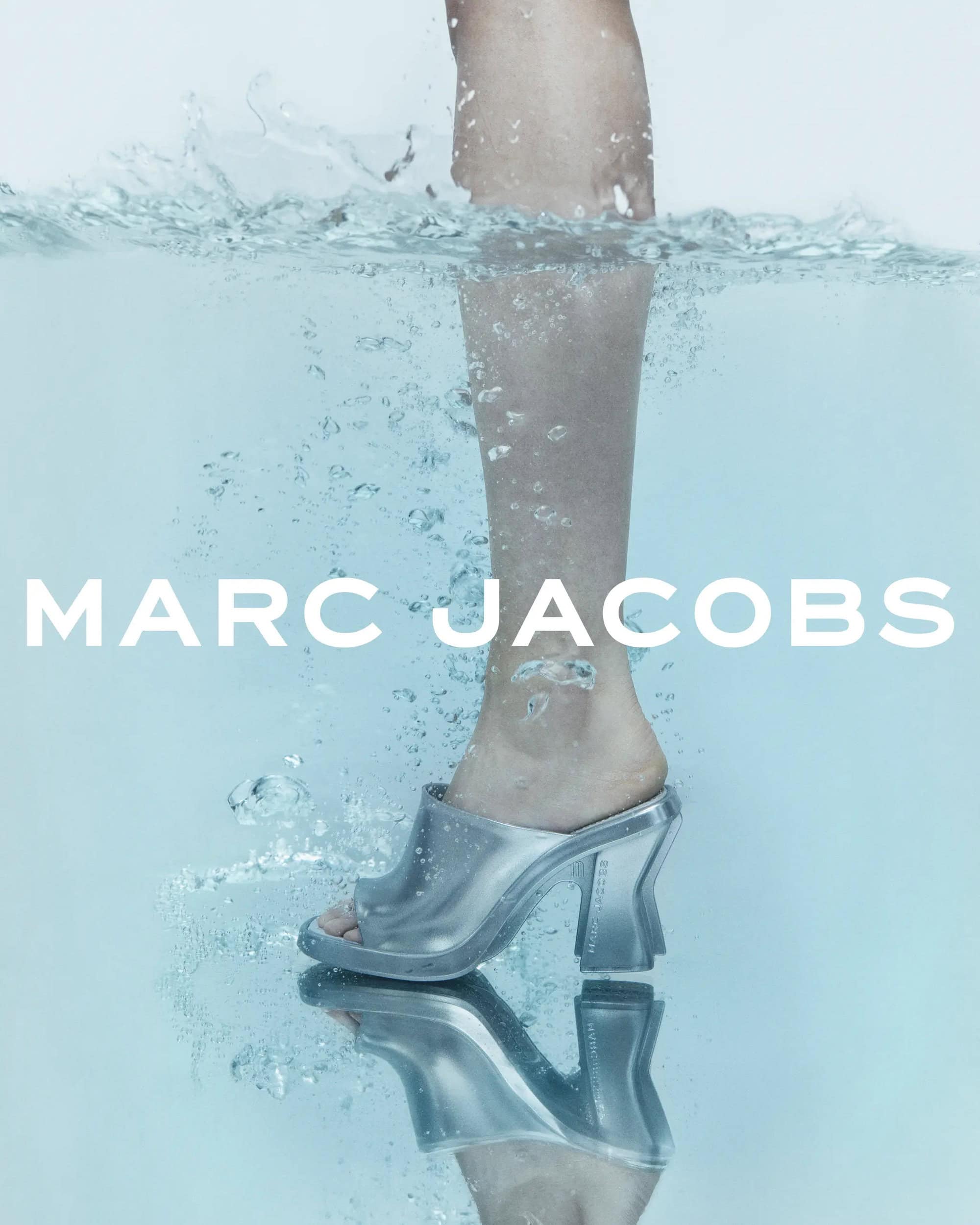 An eye-level studio shot features a leg submerged in water, wearing a translucent high-heeled shoe by Marc Jacobs, against a white backdrop.