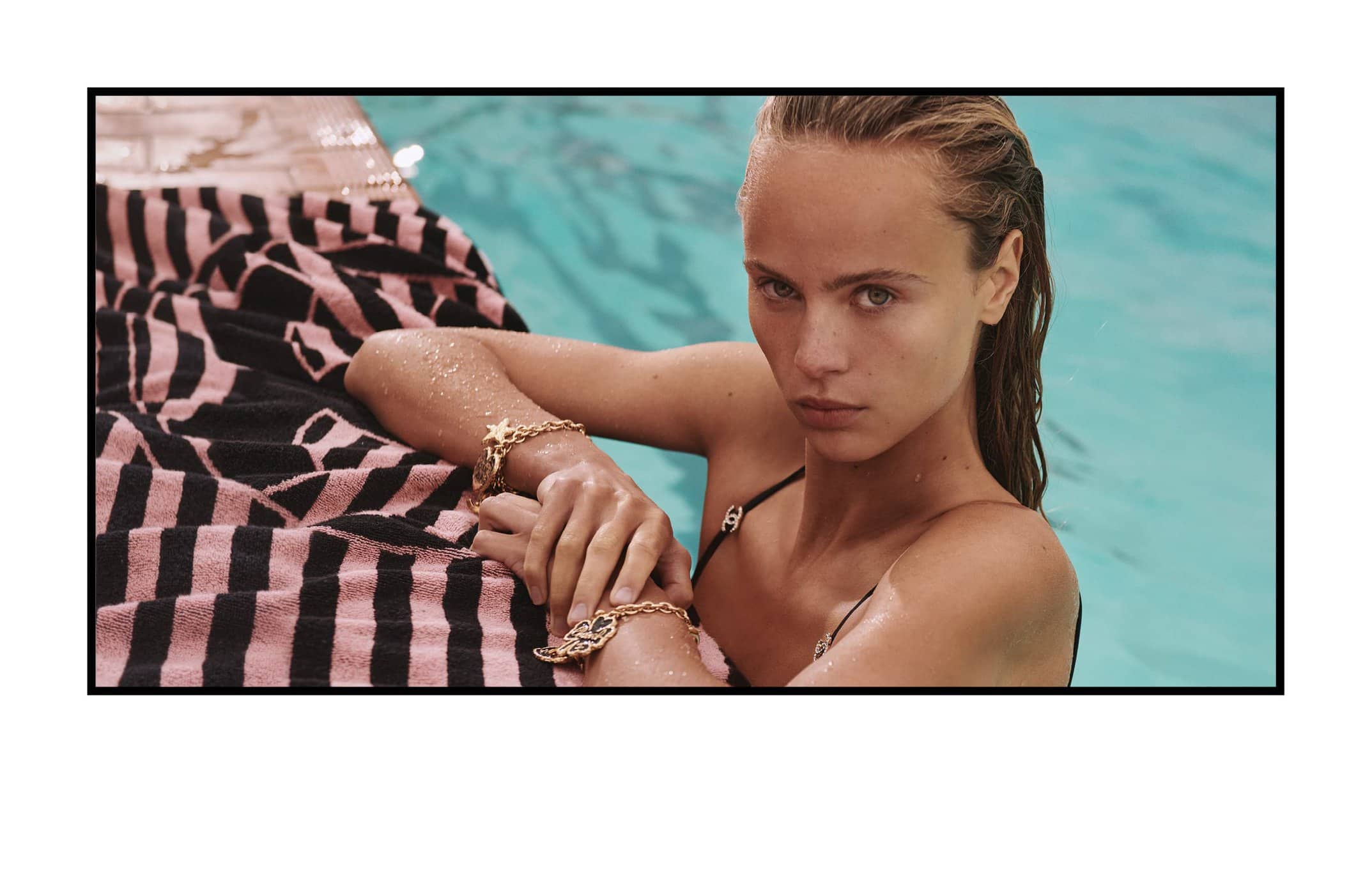 Close-up of a model with damp hair wearing gold bracelets sitting in a pool next to a pink-and-black striped towel.