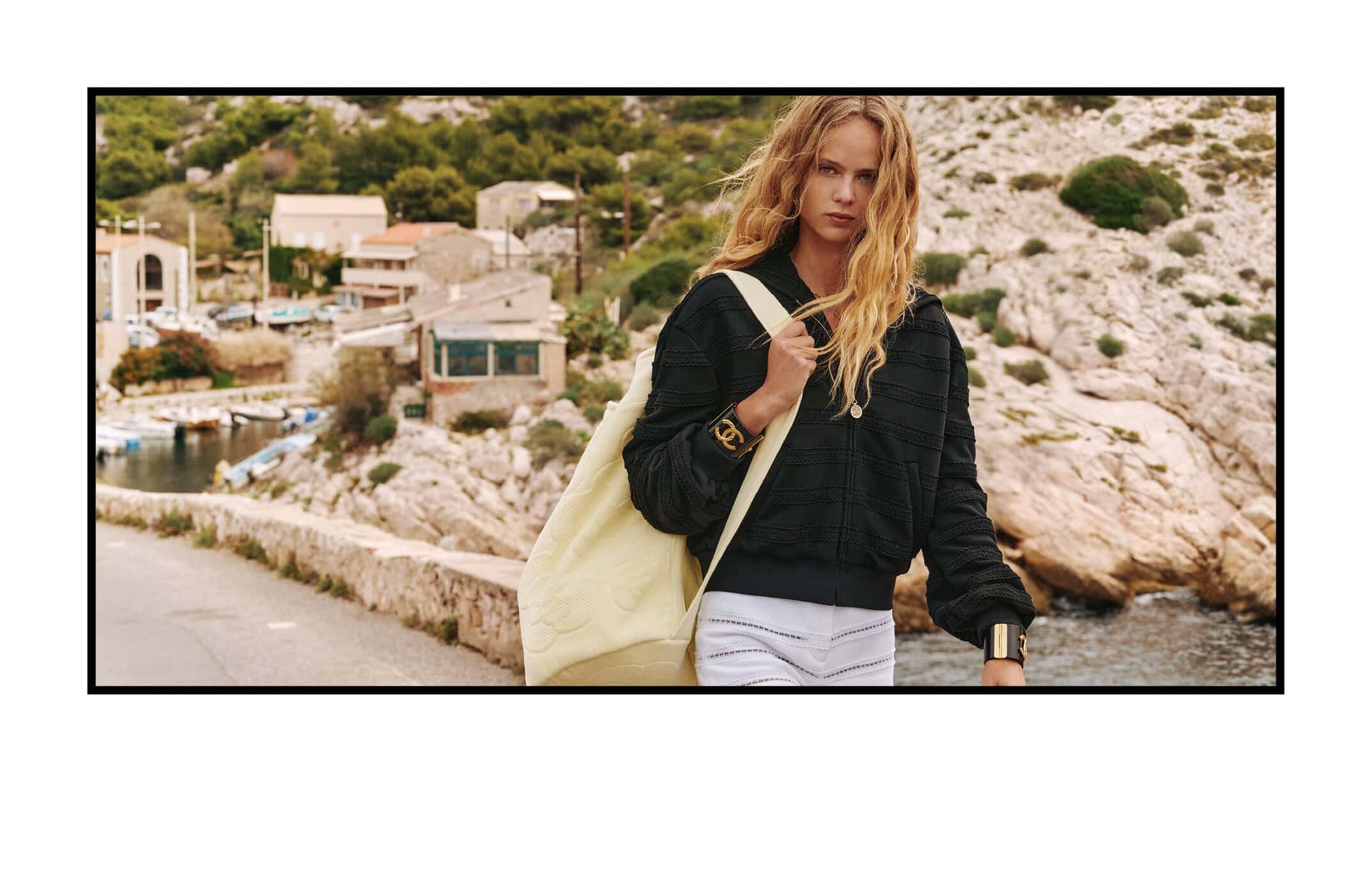 A woman poses on a stone bridge by the sea, wearing a black hooded jacket, white shorts, carrying a cream-colored tote bag.