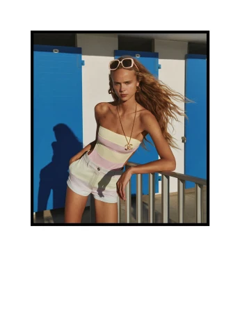 Fashion shot of a young adult female model in colorful summer clothing leaning on a railing near blue changing rooms on a sunny day.