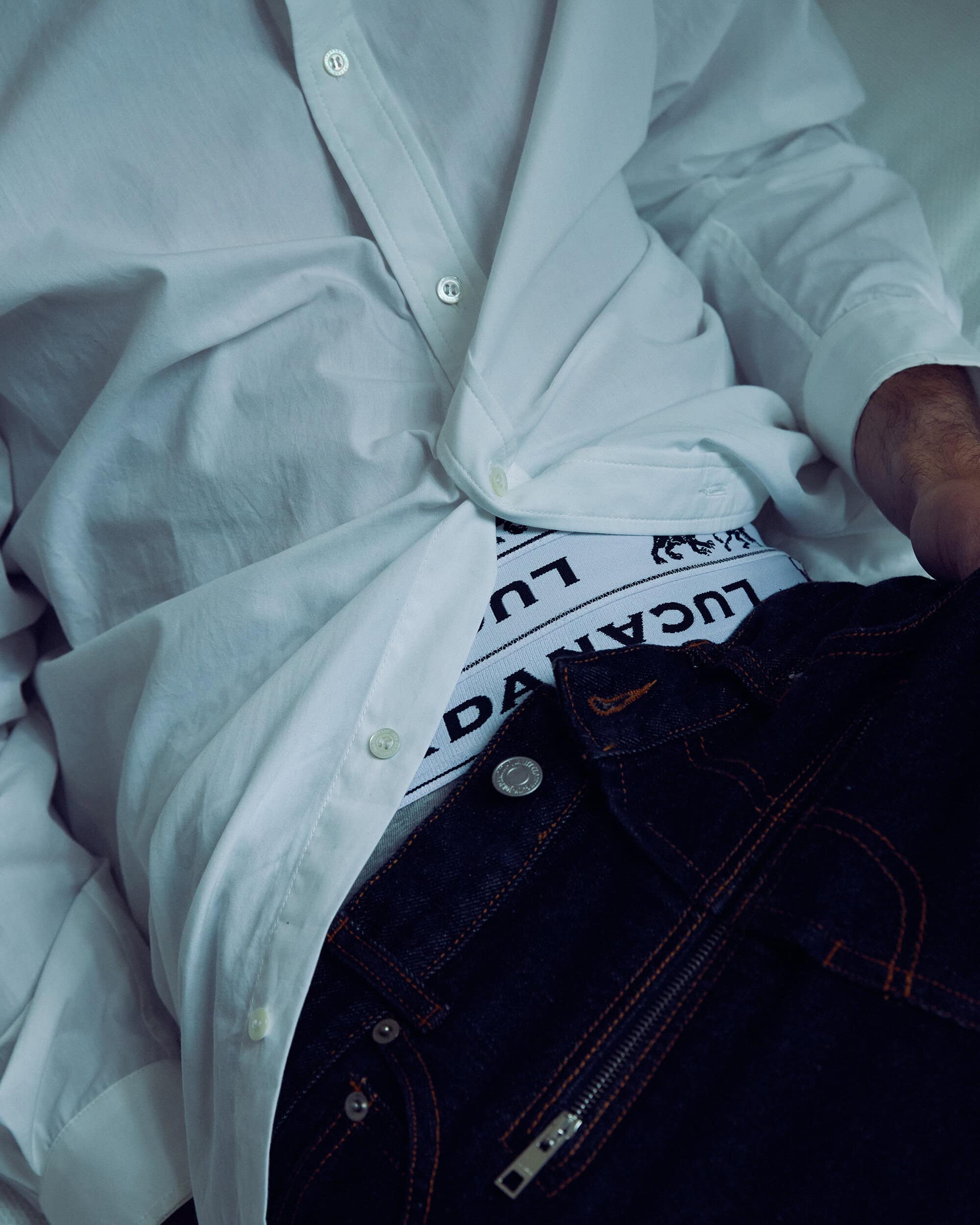 Partial view of a person reclining, wearing a white shirt, underpants with a logo, and dark jeans with zipper detailing.