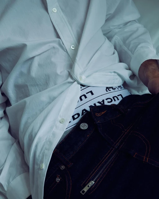 Partial view of a person reclining, wearing a white shirt, underpants with a logo, and dark jeans with zipper detailing.