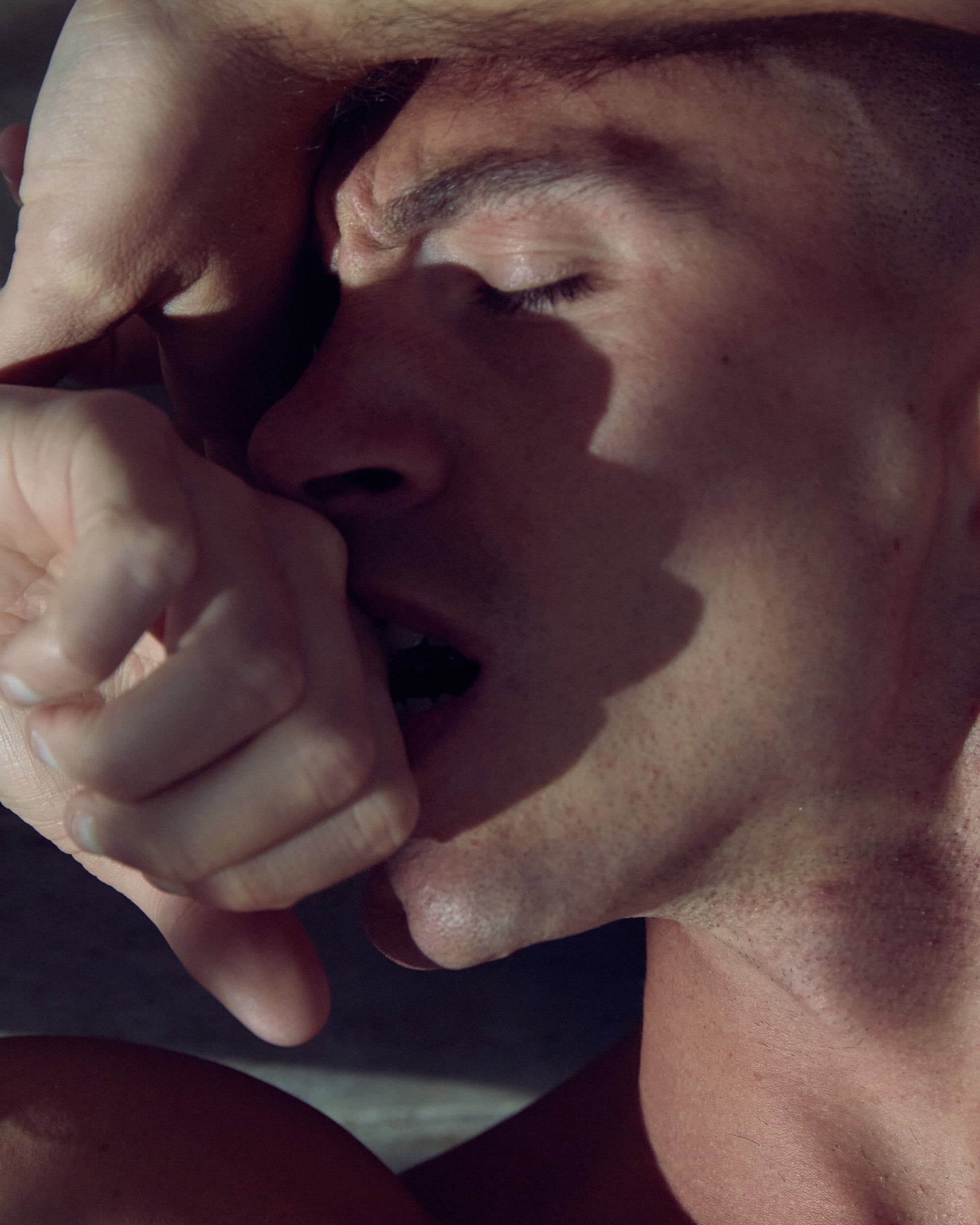 Intense close-up of a man with closed eyes, his hand near his mouth in shadow, capturing raw emotion and dramatic lighting.