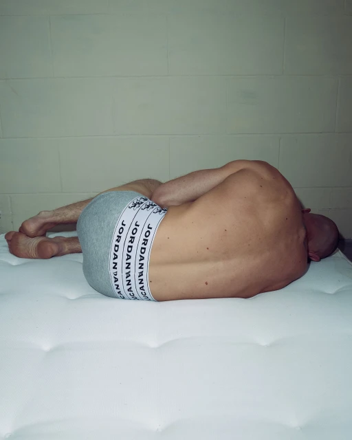 Man lying on a white mattress, wearing grey Jordan underwear, with white block wall background. The scene is softly illuminated.