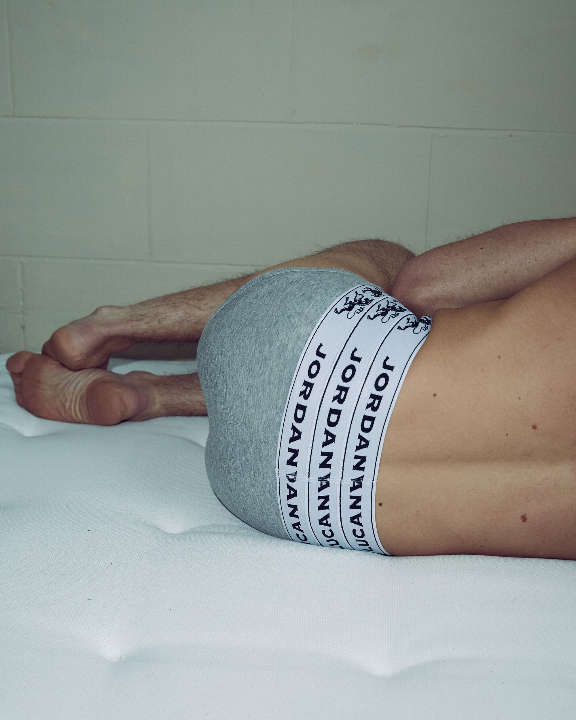 Man lying on a white mattress, wearing gray underwear with black-and-white branded waistband, and facing away from the camera.