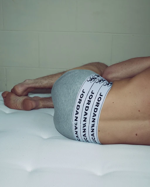Man lying on a white mattress, wearing gray underwear with black-and-white branded waistband, and facing away from the camera.