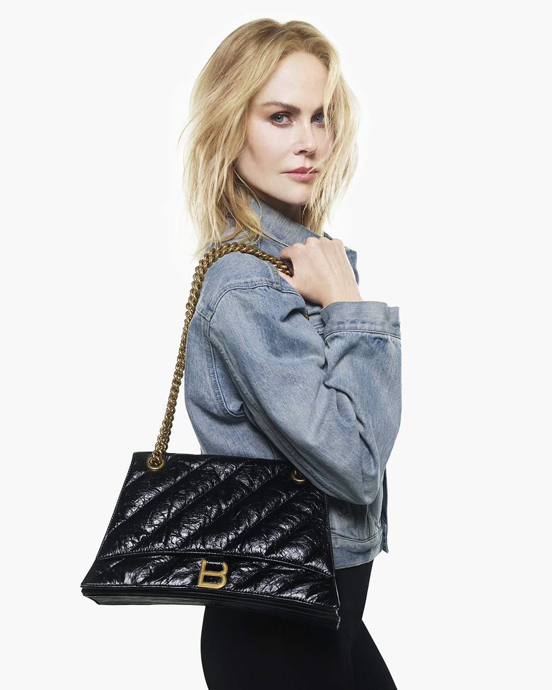 Nicole Kidman wearing denim jacket holding black crinkled leather handbag against white background
