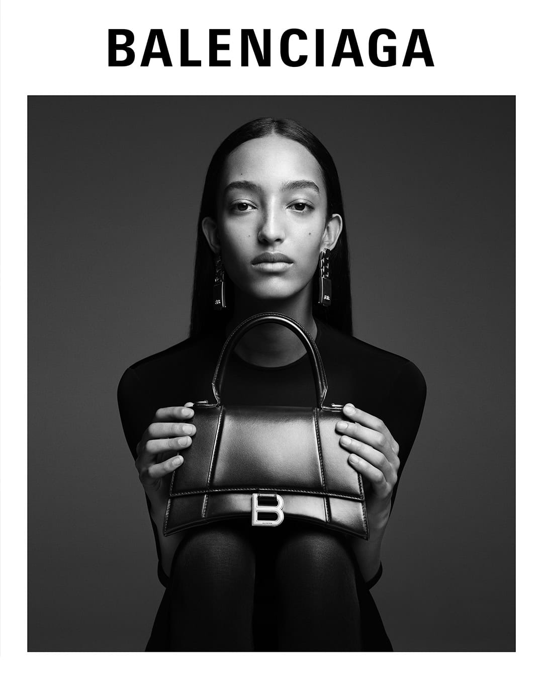 Black and white studio portrait of a model holding a structured leather handbag featuring a prominent metallic B logo