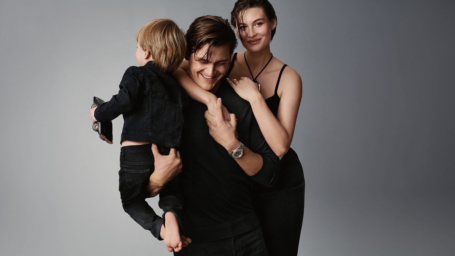Stylish family portrait featuring a father holding his young child, alongside a smiling woman, all in dark attire against a grey background.