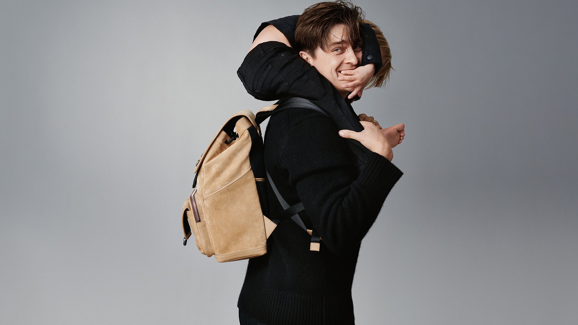 A man carries a child on his shoulders with a beige backpack, showcasing an indoor studio shot with a clean grey backdrop.