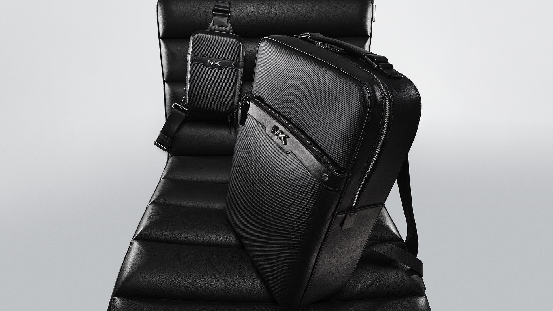 Elevated view of a black leather backpack and crossbody bag with metal logo on a black leather chair against a light gray backdrop.