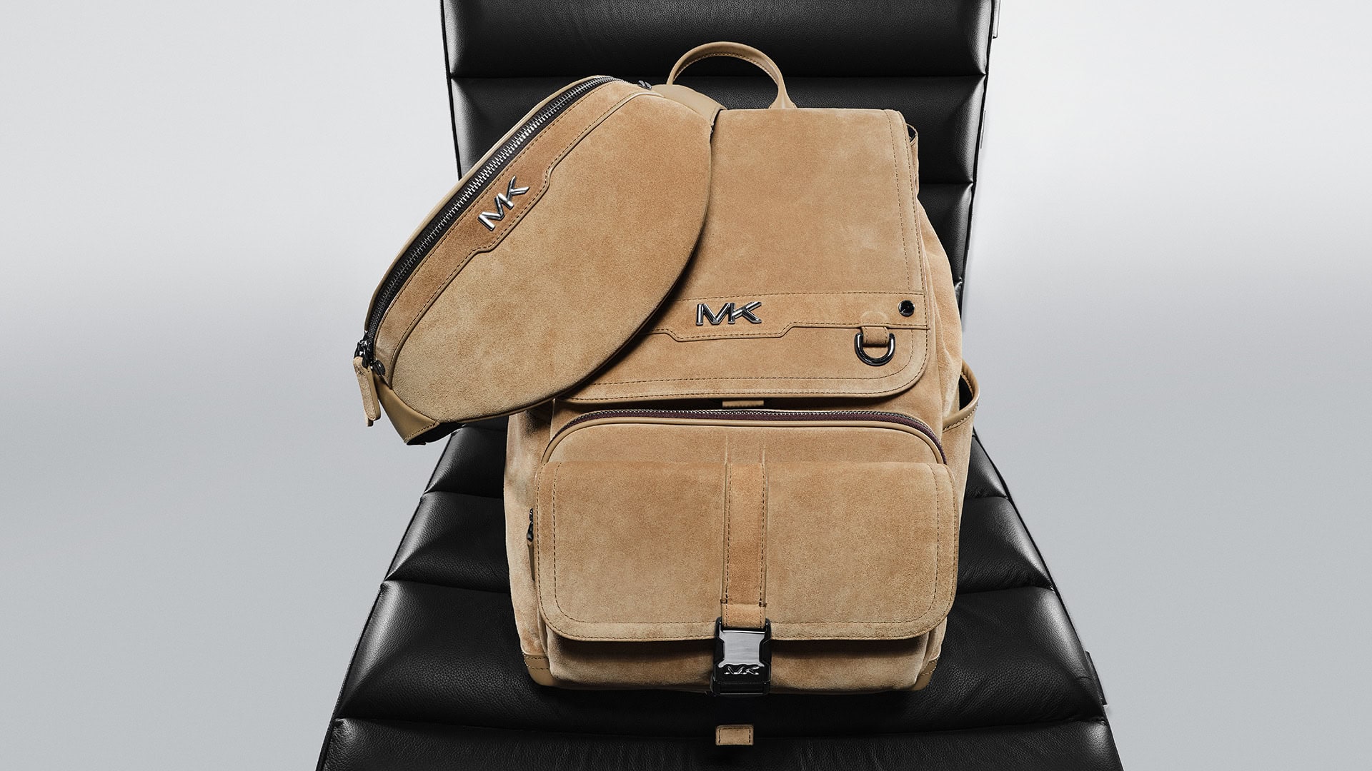 Suede backpack and belt bag with brand logos, set against a textured dark gray chair and a light gray studio background.