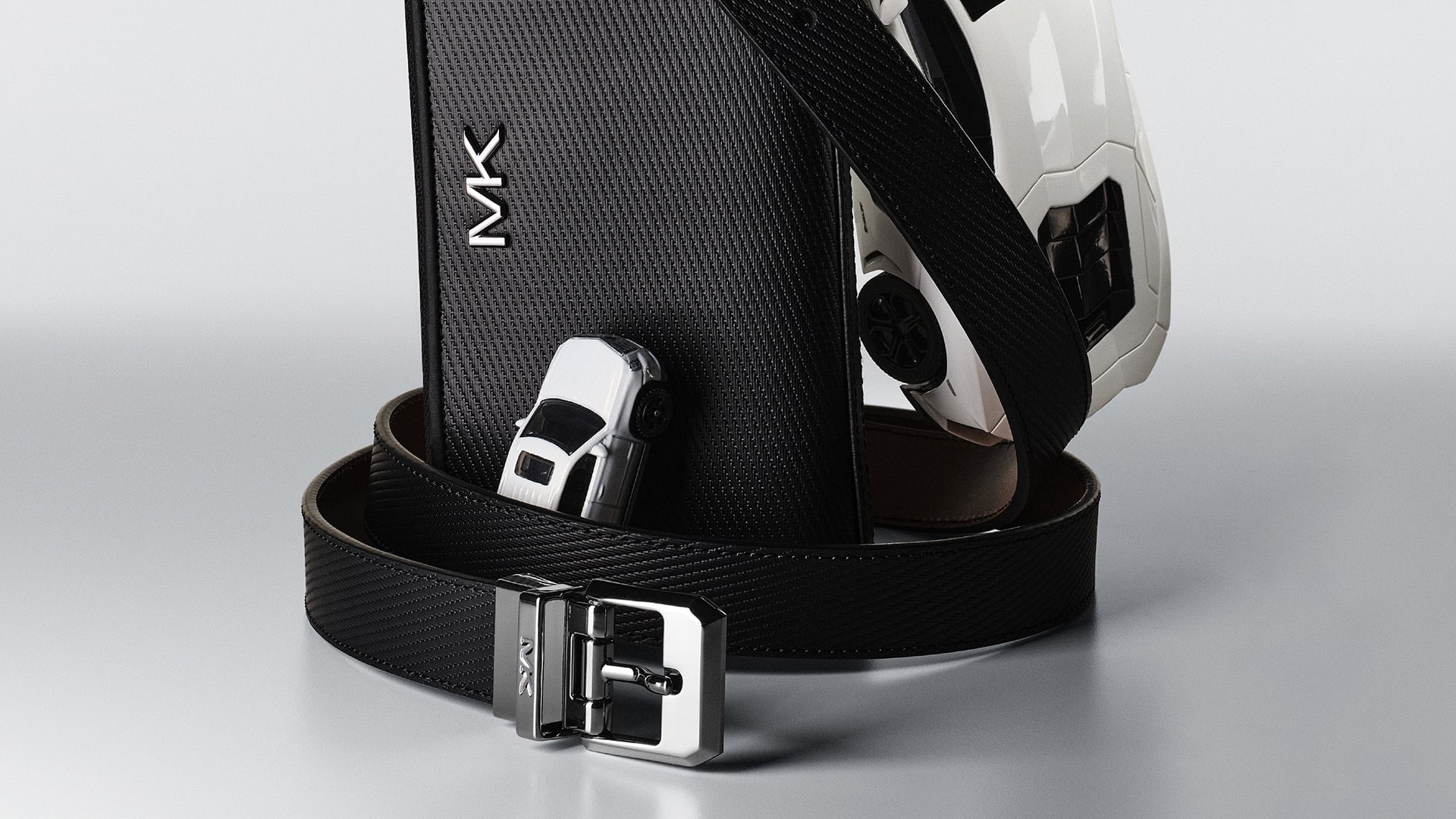 Elegant arrangement of a black textured wallet, black leather belt, and white toy sports cars on a graduated gray background.