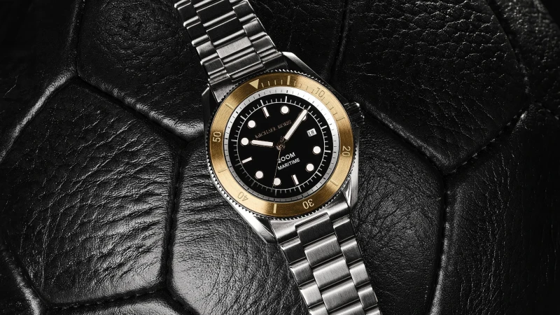 Wristwatch on a textured surface shows a stainless steel band, gold bezel, black dial, and date window for a luxury aesthetic.